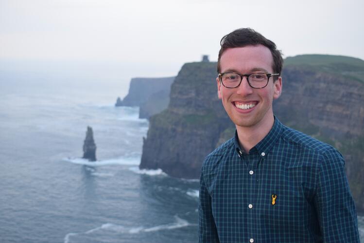 A man wearing glasses smiles at the viewer. In the background, bluffs and an ocean are visible.