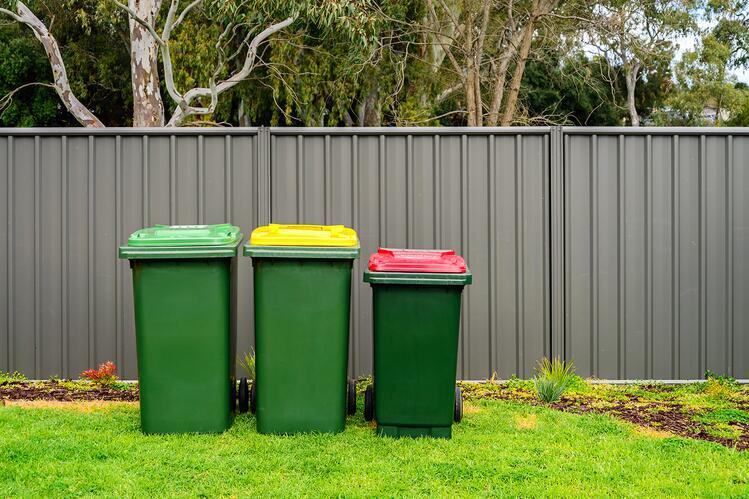three waste bins with green, yellow and red lids placed in a yard against a fence.