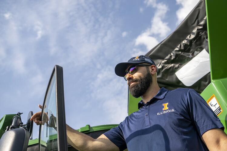 Man with sunglasses and Illinois hat and t-shirt poses by a tractor with blue sky in the background. 